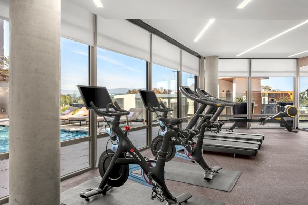 The image shows a modern gym with exercise bikes and treadmills beside large windows overlooking an outdoor pool terrace.