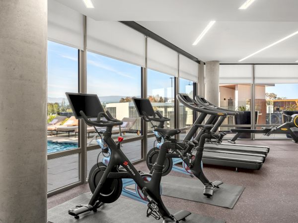 The image shows a modern gym with exercise bikes and treadmills beside large windows overlooking an outdoor pool terrace.