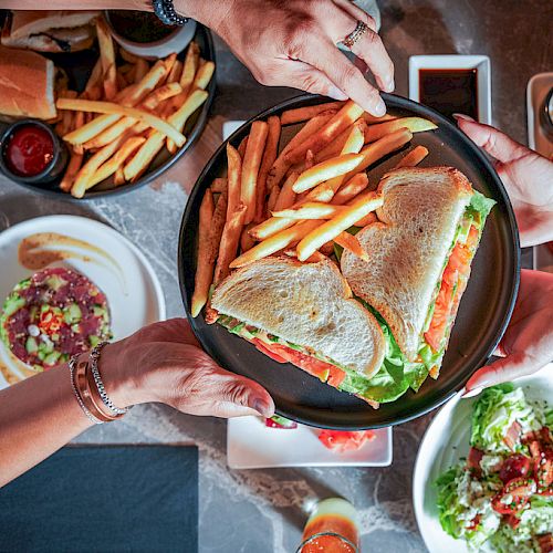 Friends share a platter of sandwiches and fries at a table, reaching for food in a casual meal.