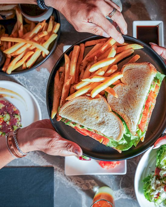 Friends share a platter of sandwiches and fries at a table, reaching for food in a casual meal.