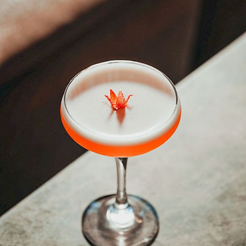 A white cocktail with foamy top and a small red flower garnish, served in a glass with an orange rim.