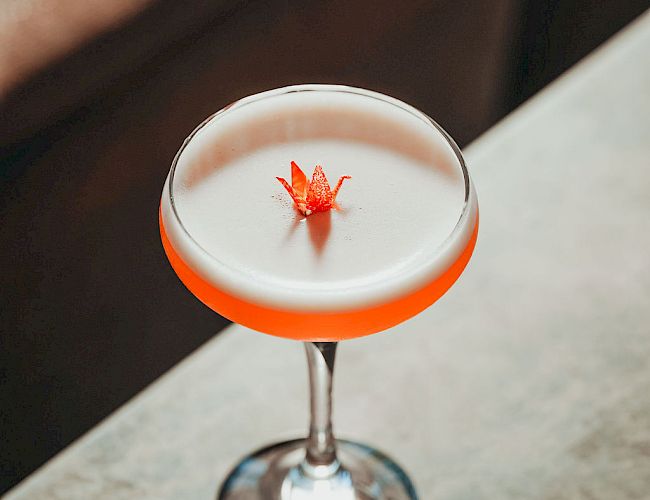 A white cocktail with foamy top and a small red flower garnish, served in a glass with an orange rim.