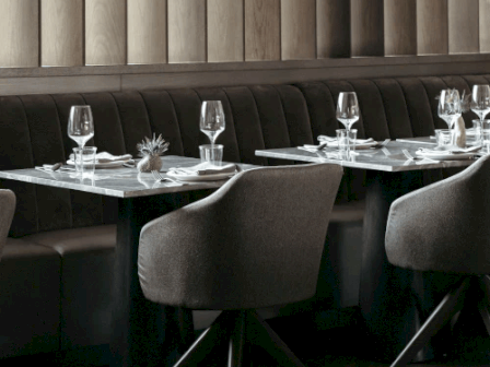 A modern restaurant booth with dark banquettes, set dining tables, glassware, and a pine-wood paneled wall.