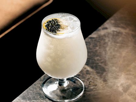 A frothy white cocktail in a stemmed glass, topped with black garnish, on a marble table with warm bar lighting.