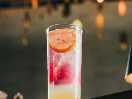 A tall glass cocktail with red and pale layers, chilled with ice, shot against a blurry bar background.