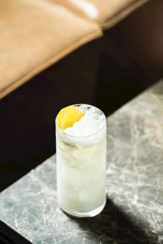 A tall iced drink in a glass with carbonation and a lemon slice garnish, sitting on a marble bar counter.
