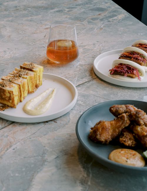 Plates with stuffed buns, fried food, a slice dish with sauce, and a glass of a drink are on a table.