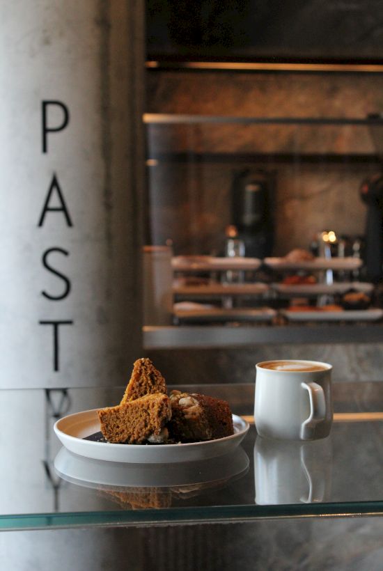A slice of cake on a plate and a cup of coffee are placed on a glass table in a pastry shop.