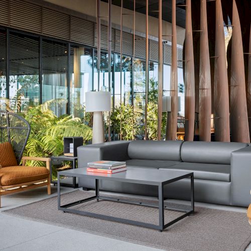 A modern outdoor seating area with a gray sofa, two armchairs, coffee table, and decorative plants, set against glass walls and wooden decor.