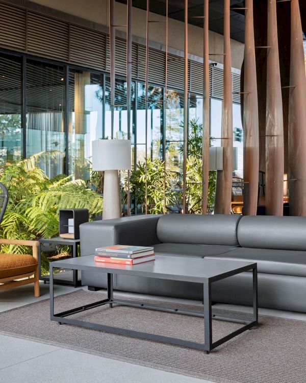 A modern outdoor seating area with a gray sofa, two armchairs, coffee table, and decorative plants, set against glass walls and wooden decor.