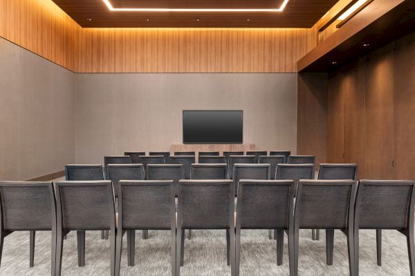 The image shows a small conference room with rows of chairs facing a large screen on the wall, with wood paneling and carpeted flooring.