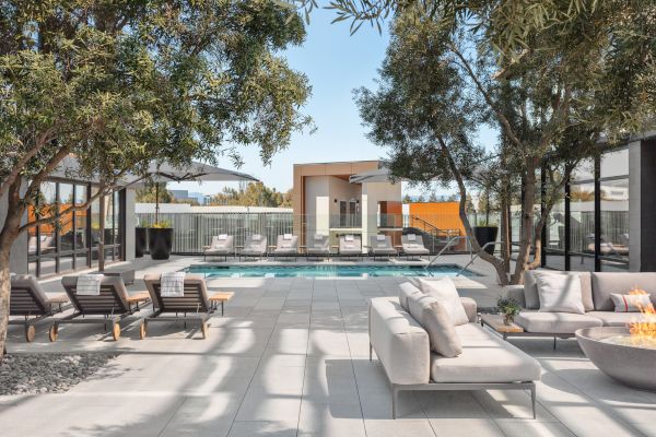 The image shows a modern outdoor space with a swimming pool, lounge chairs, sofas, trees, and a fire pit, surrounded by glass walls.