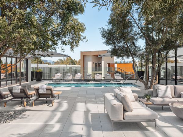 The image shows a modern outdoor space with a swimming pool, lounge chairs, sofas, trees, and a fire pit, surrounded by glass walls.