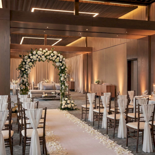 An elegant wedding venue: rows of white-chair covers, aisle petals, and a floral arch altar under warm lighting.