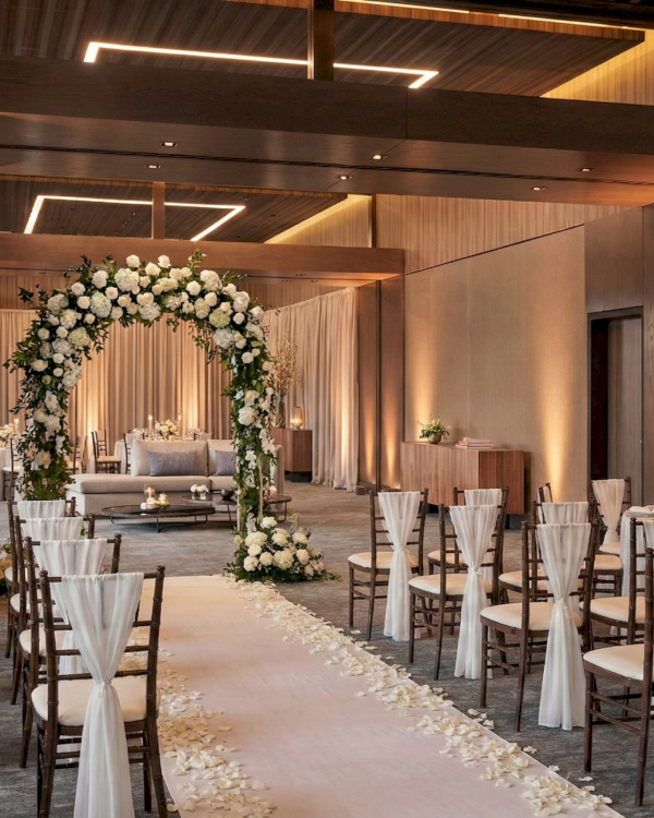 An elegant wedding venue: rows of white-chair covers, aisle petals, and a floral arch altar under warm lighting.