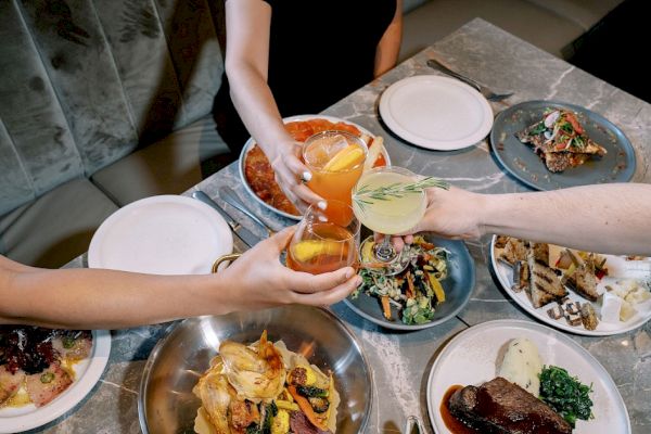 People are toasting with drinks over a table filled with various plates of food, including meat dishes and salads.