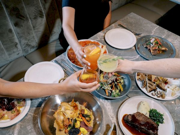People are toasting with drinks over a table filled with various plates of food, including meat dishes and salads.