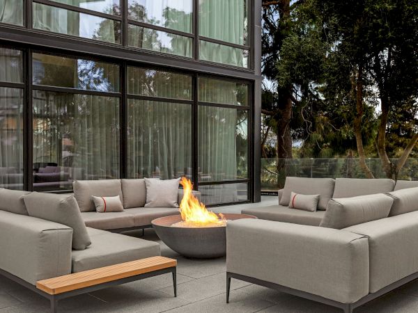A modern outdoor seating area with gray sofas surrounds a lit fire pit, positioned near a building with large glass windows.