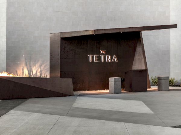 A modern entrance with "TETRA" displayed on a dark panel, flame feature to the left, and a minimalist design in a gray setting.