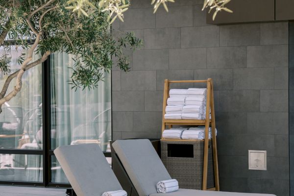 Two lounge chairs with rolled towels are placed under a tree near a modern building.