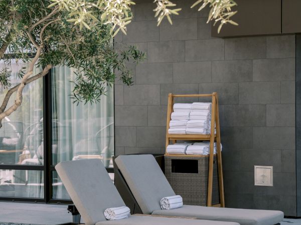 Two lounge chairs with rolled towels are placed under a tree near a modern building.