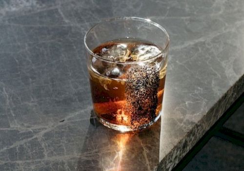 A glass of a dark beverage with ice sits on a marble surface, casting shadows and highlights from the light.