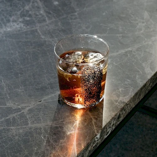 A glass of a dark beverage with ice sits on a marble surface, casting shadows and highlights from the light.