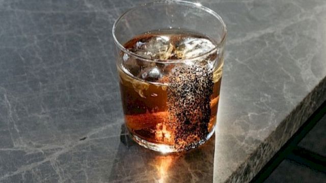 A glass of a dark beverage with ice sits on a marble surface, casting shadows and highlights from the light.