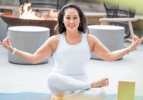A person is meditating in a cross-legged pose on a yoga mat, with a fire pit and chairs in the background.