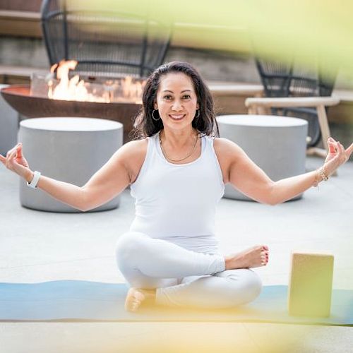 A person is meditating in a cross-legged pose on a yoga mat, with a fire pit and chairs in the background.