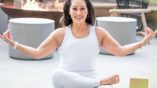 A person is meditating in a cross-legged pose on a yoga mat, with a fire pit and chairs in the background.