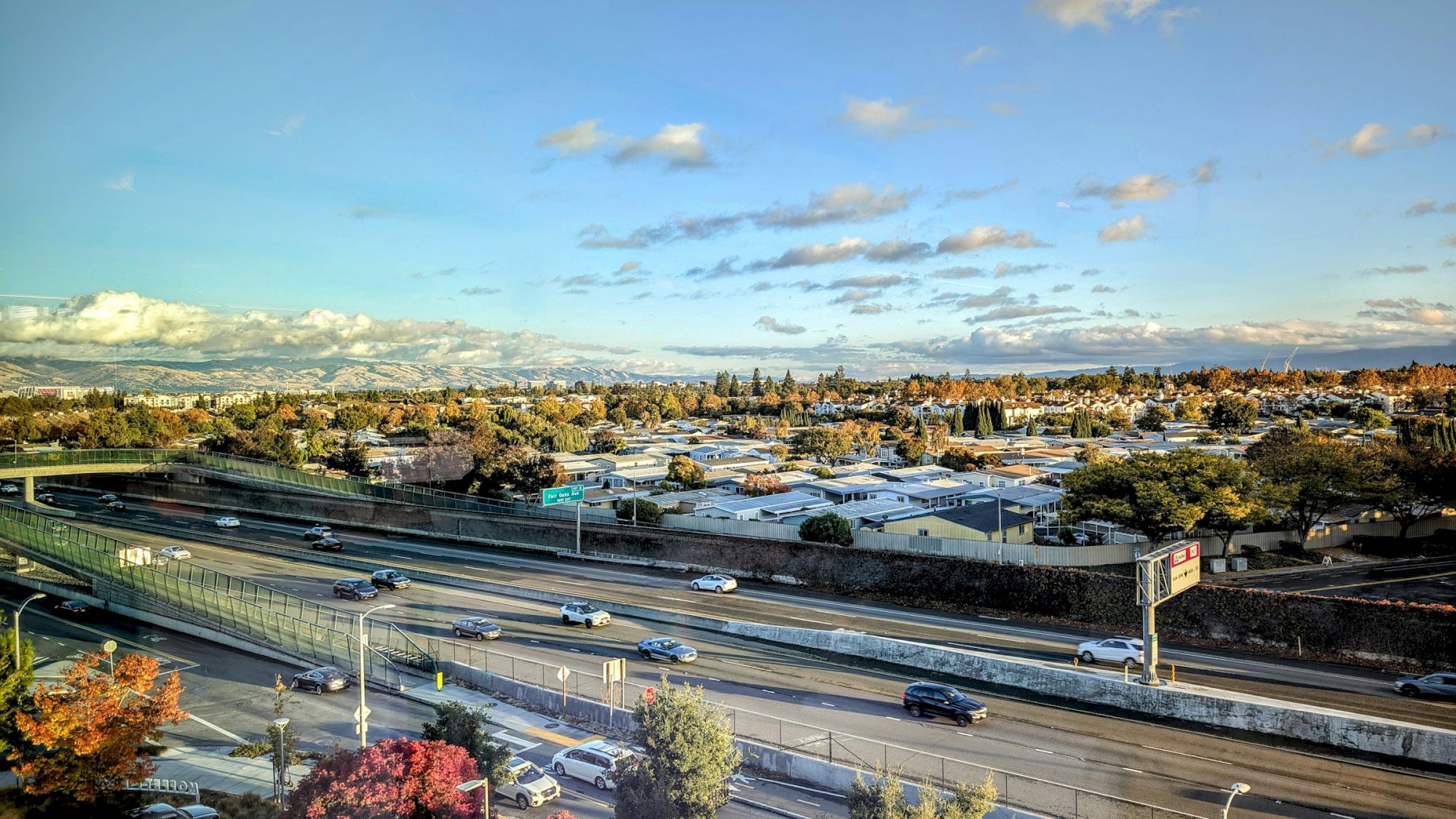 A highway with moving cars, surrounded by trees and buildings under a blue, partly cloudy sky.