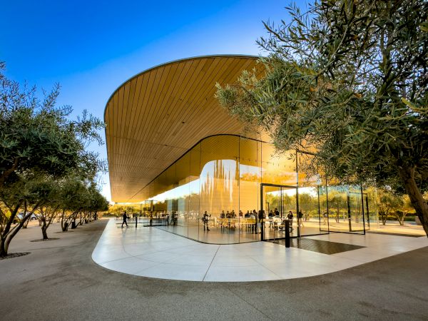 A modern building with curved glass walls and a sleek wooden roof, surrounded by trees and people gathered around it.