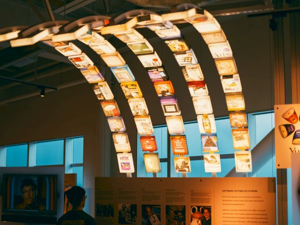 The image shows a display with various illuminated hanging panels, each showing different designs and images, inside a modern exhibition space.