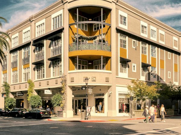 A multi-story building with shops at street level, located on a sunny corner with people walking by and cars parked nearby.