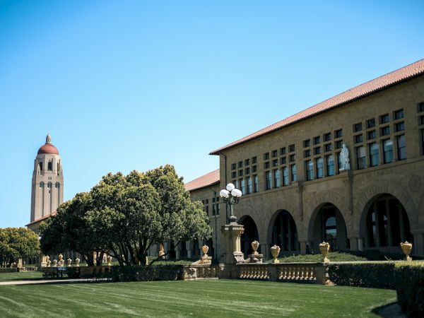 A grassy lawn leads to a large building with arched windows and a red-roofed tower against a clear blue sky.