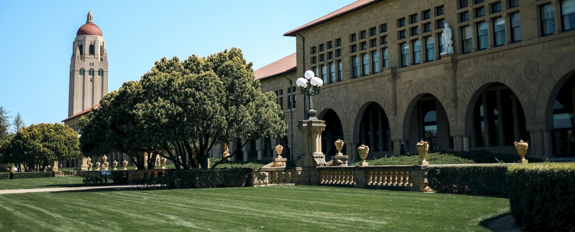 A large building with arched windows, a tall tower, and trees on a well-maintained lawn under a clear blue sky.