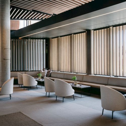 The image shows a modern lounge area with chairs, tables, and vertical blinds on windows, creating a cozy setting with natural light.
