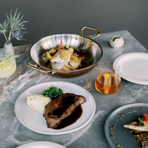 A table setting with plated meats, mashed potatoes, a pan with garnish, cocktails, and minimalistic decor, over a gray surface.