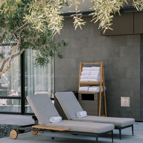 Two lounge chairs with towels beside a tree, next to a modern building and towel rack, creating a serene outdoor setting.