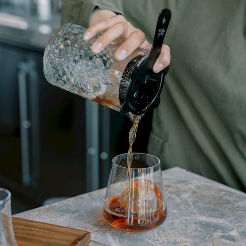 A person pours a cold brew coffee from a jar into a glass with ice on a table.