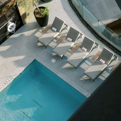 A rooftop pool with five lounge chairs and potted plants, surrounded by glass railings and shadows cast by nearby structures.