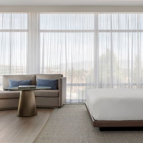 The image shows a modern hotel room with a bed, sofa, desk, round table, and large windows with sheer curtains.