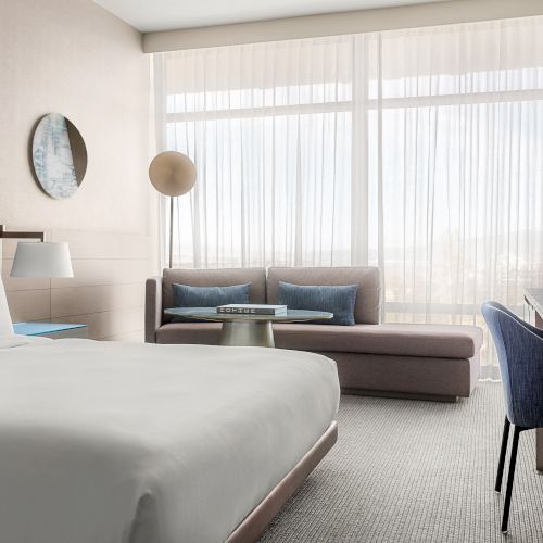 A modern hotel room with a large bed, sofa, desk, and floor-to-ceiling windows with sheer curtains, creating a bright and inviting space.