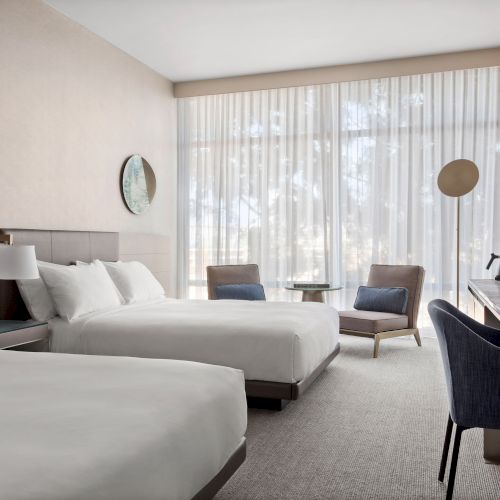 The image shows a modern hotel room with two beds, a desk, chairs, and a large window with sheer curtains, creating a cozy atmosphere.