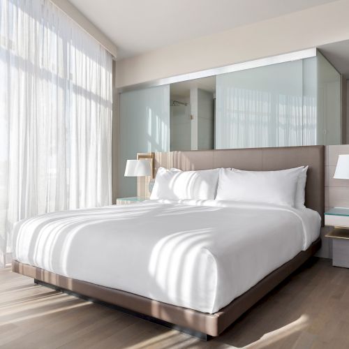 A modern hotel room with a large bed, white bedding, bedside lamps, and floor-to-ceiling windows with sheer curtains.