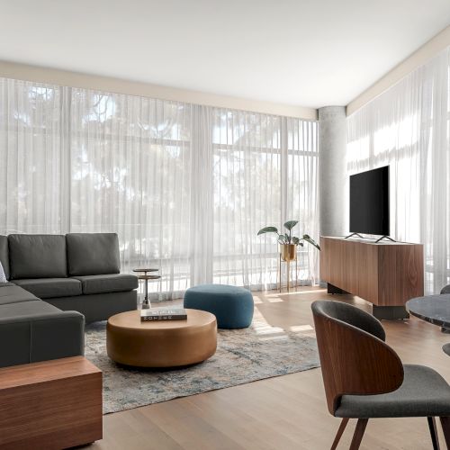 A modern living room with a sectional sofa, TV on a console, round coffee table, chairs, and a dining table, all in natural light.