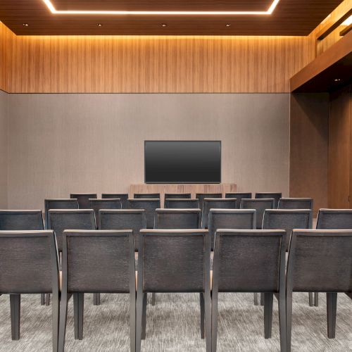 The image shows a modern conference room with rows of chairs facing a flat-screen TV on the front wall.