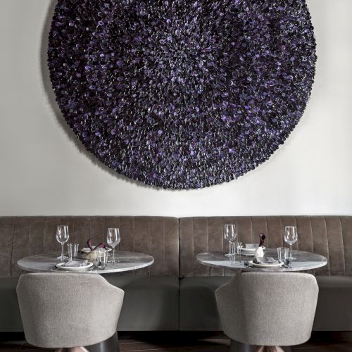 Two small tables with chairs, set for dining, are placed beneath a large circular textured artwork on the wall.