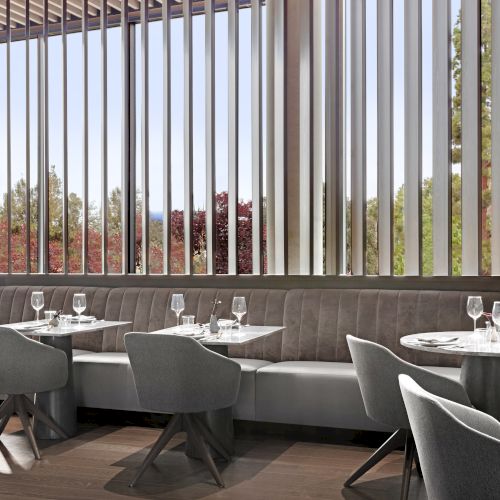 A modern restaurant interior with tables, gray chairs, set with glasses and napkins, features vertical slats with outdoor views.